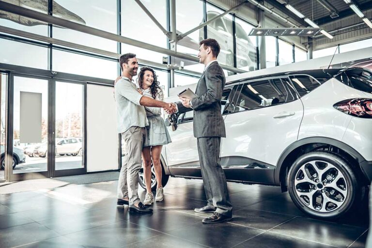 A person reviews a price comparison printout with a salesperson next to a parked vehicle in a dealership lot.